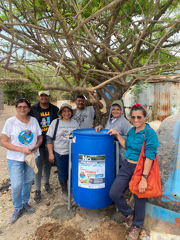 a group of smiling people around a big blue trash can under a tree in El Salvador as part of an effort to keep trash out of the coastal environment.