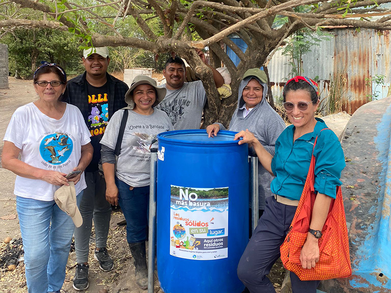Un grupo de personas sonrientes alrededor de un gran bote de basura azul debajo de un árbol, como parte de un esfuerzo por mantener la basura fuera del ambiente costero en El Salvador.