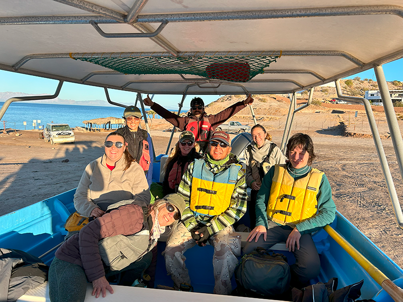 a group of eight people smiling and sitting in a boat on the shore
